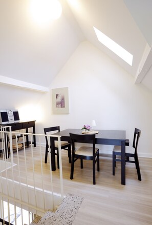 A modern, well-lit dining area with a high ceiling and skylight, featuring a dark wooden table and chairs, a small side table with a lamp, and a laptop on a desk. - Bauerncafé & Pension Drostenhof (Rees)