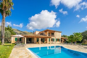 Una casa de piedra de dos pisos con piscina, mobiliario exterior y el paisaje de montañas al fondo.