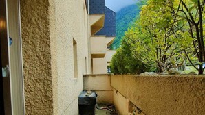 A narrow outdoor corridor with a textured beige wall, a small step leading to a door, and a tree with green leaves to the right.
