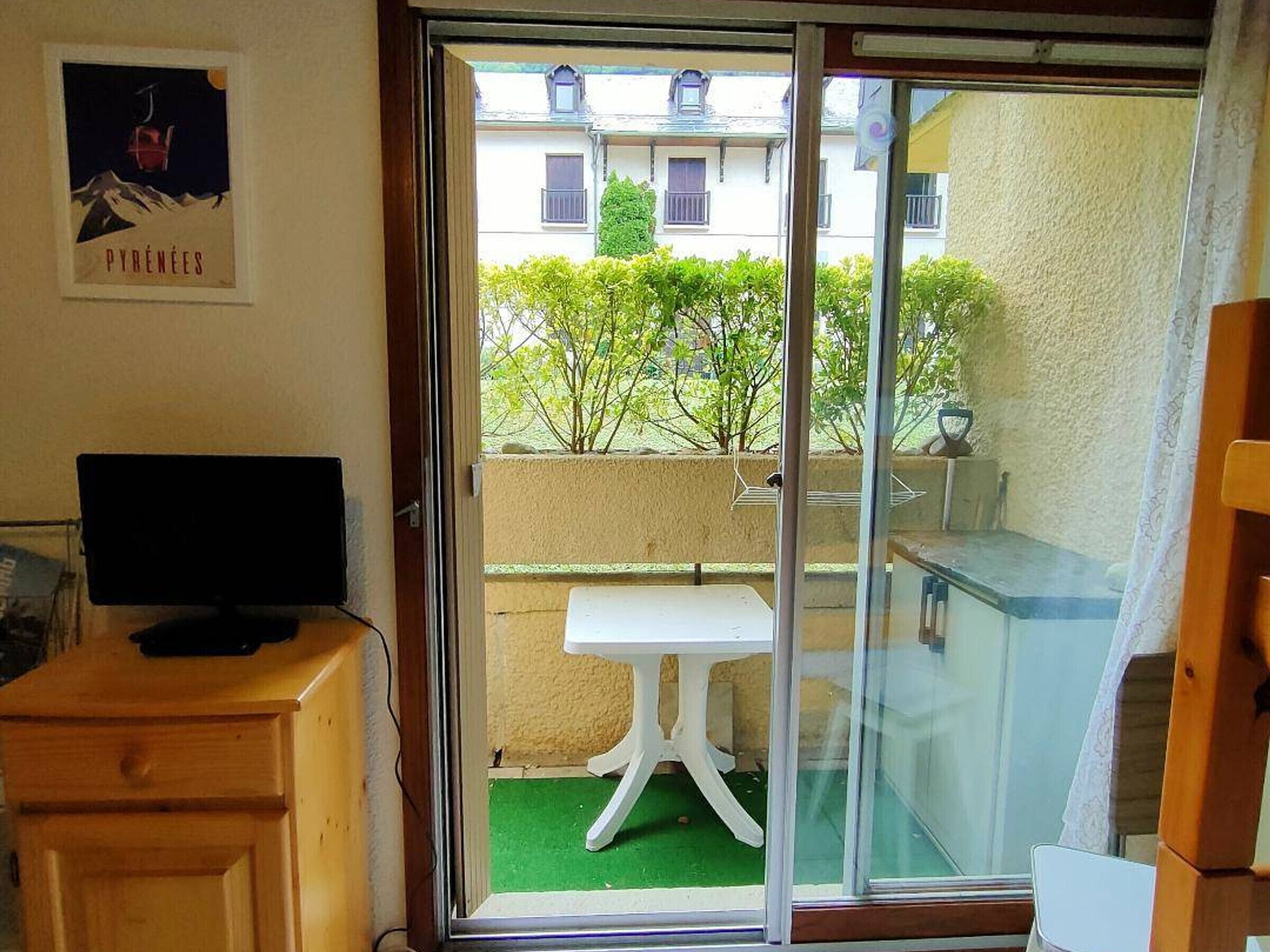 A room with a sliding glass door leading to a balcony, a small white table, and a wooden cabinet with a television.