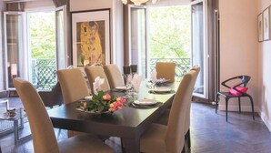 A dining area with a long table set for six, large windows with a view of greenery, and a chandelier. - Enjoy Eiffel Tower Sparkle in Grand Style! (Paris)