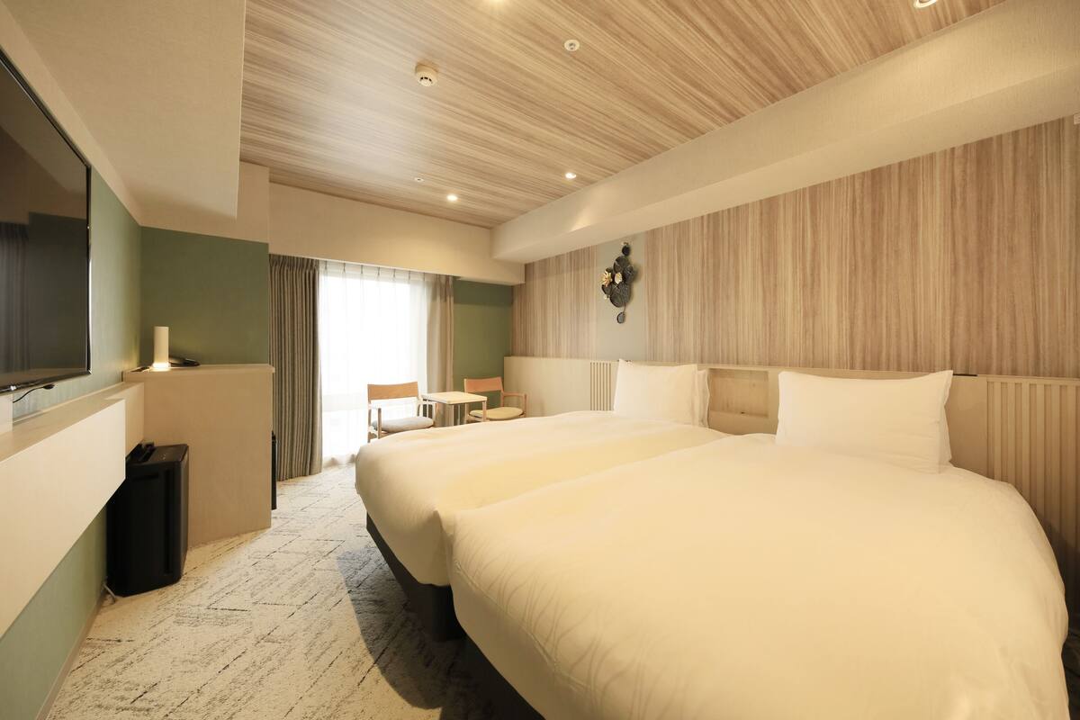 A hotel room with two large beds, a small table, a chair, a TV, and a waste bin.
