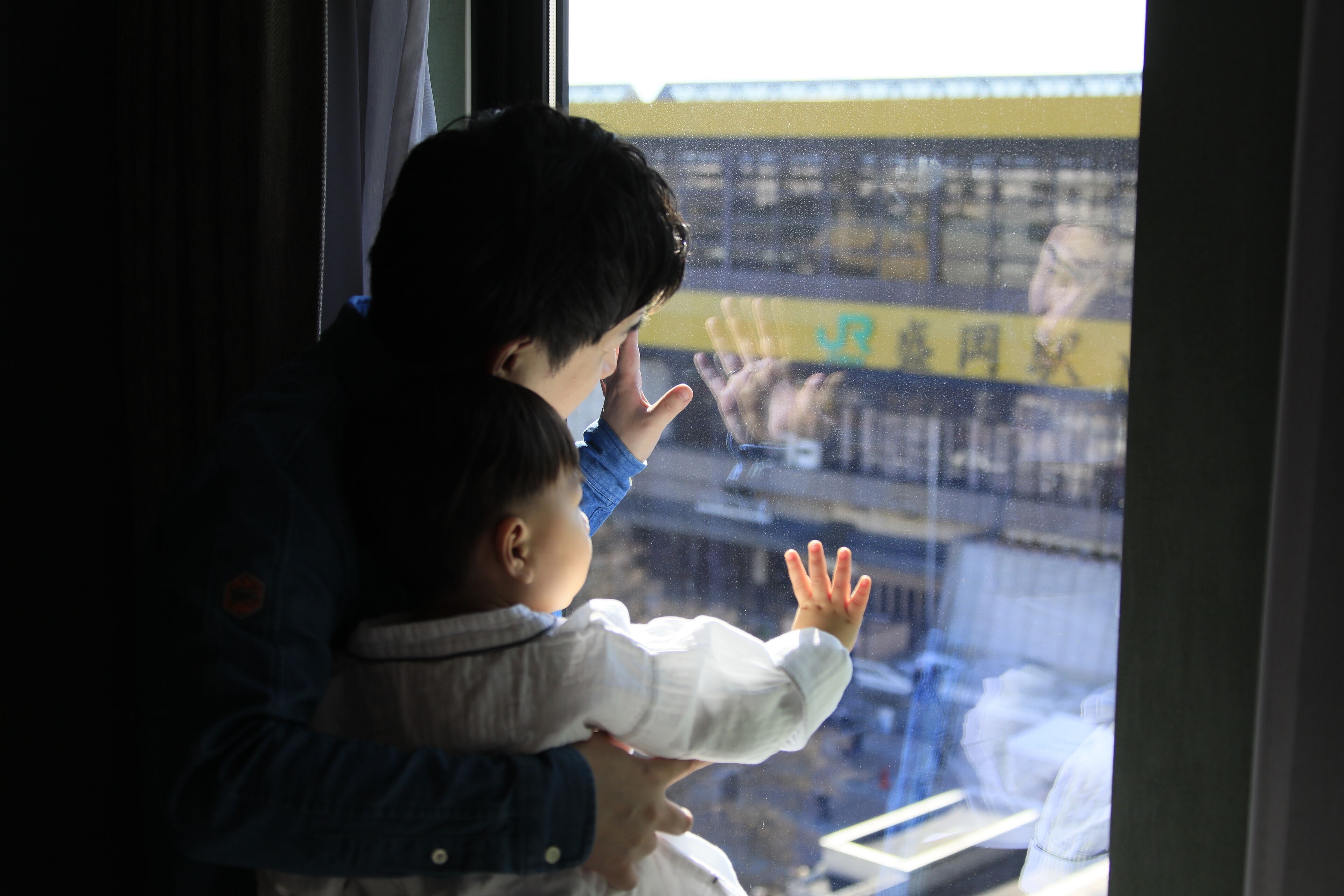 Una persona sostiene a un niño junto a una ventana con vistas a un edificio con señalización amarilla y azul.