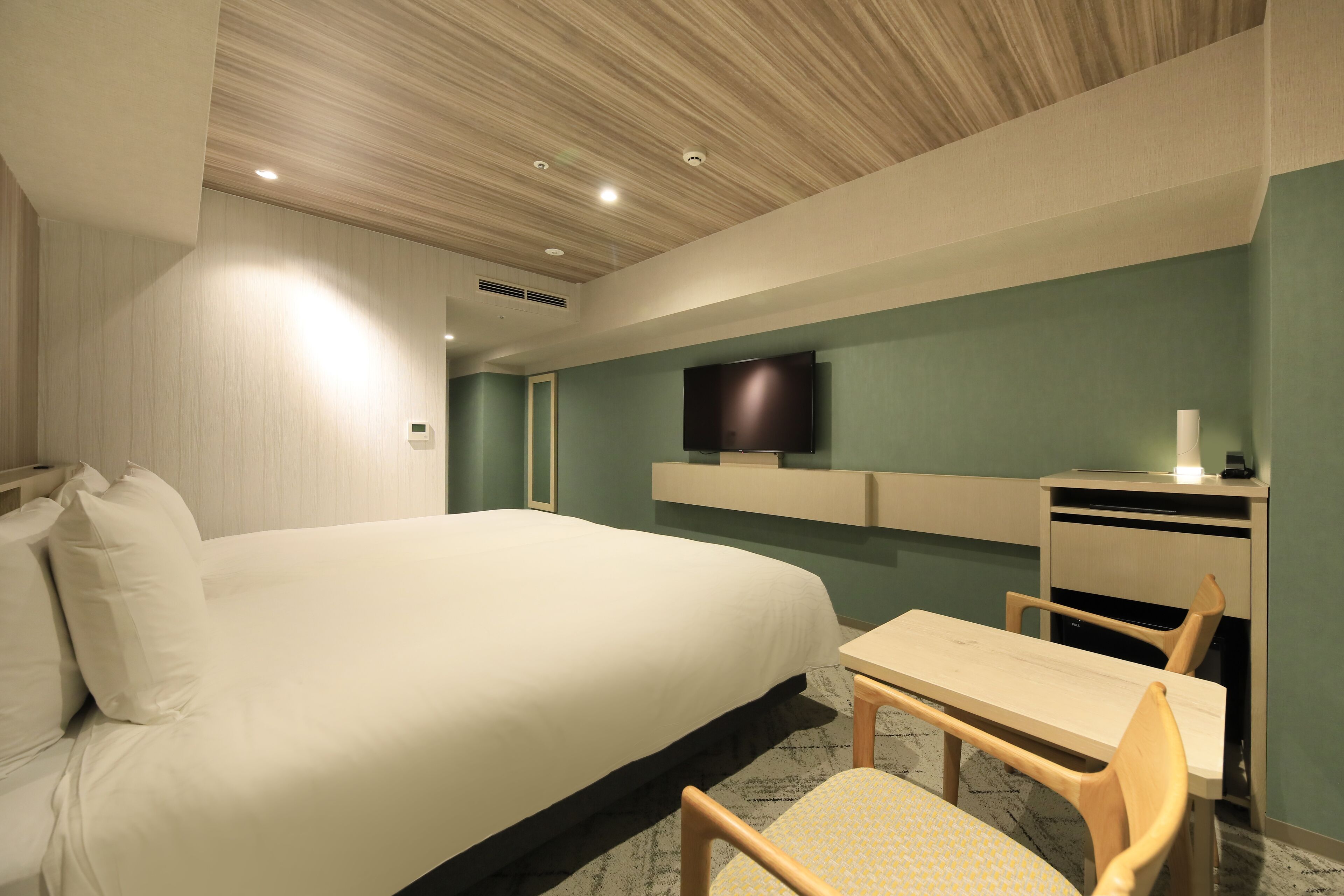 A modern hotel room with a large bed, a desk with a chair, a flat-screen TV mounted on the wall, and a small table.