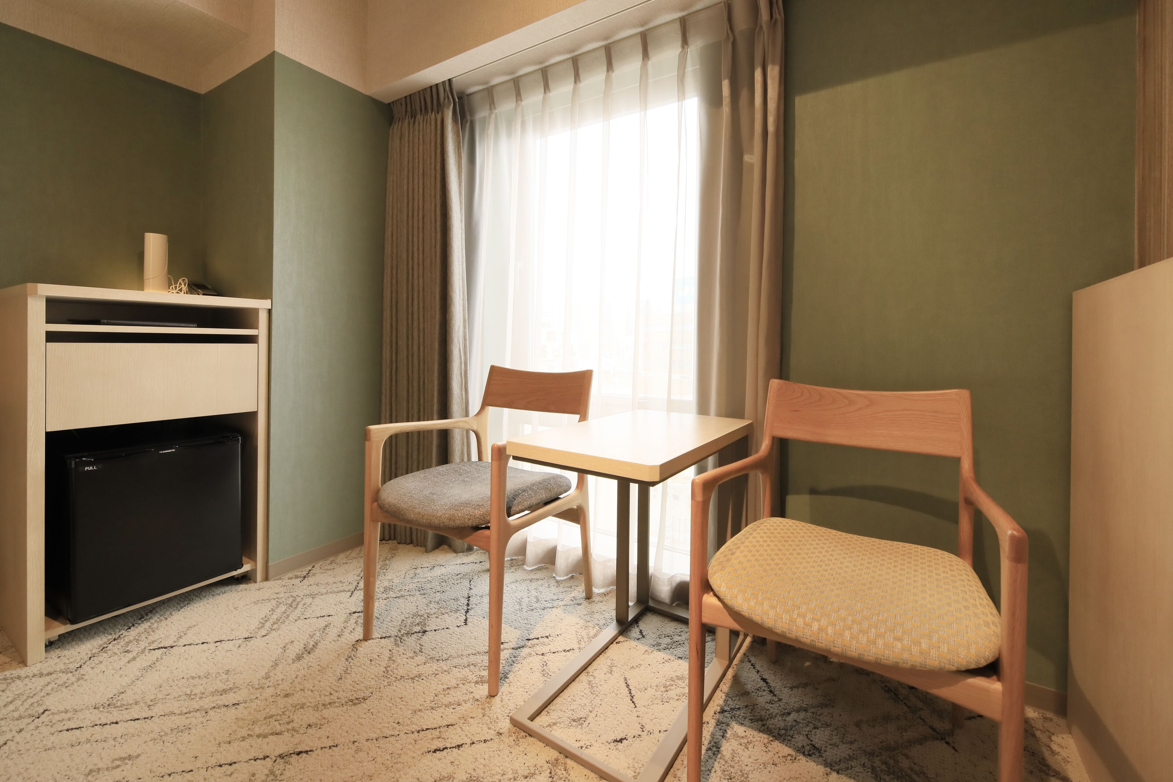 A small room with a desk, two chairs, a refrigerator, and a window with curtains.