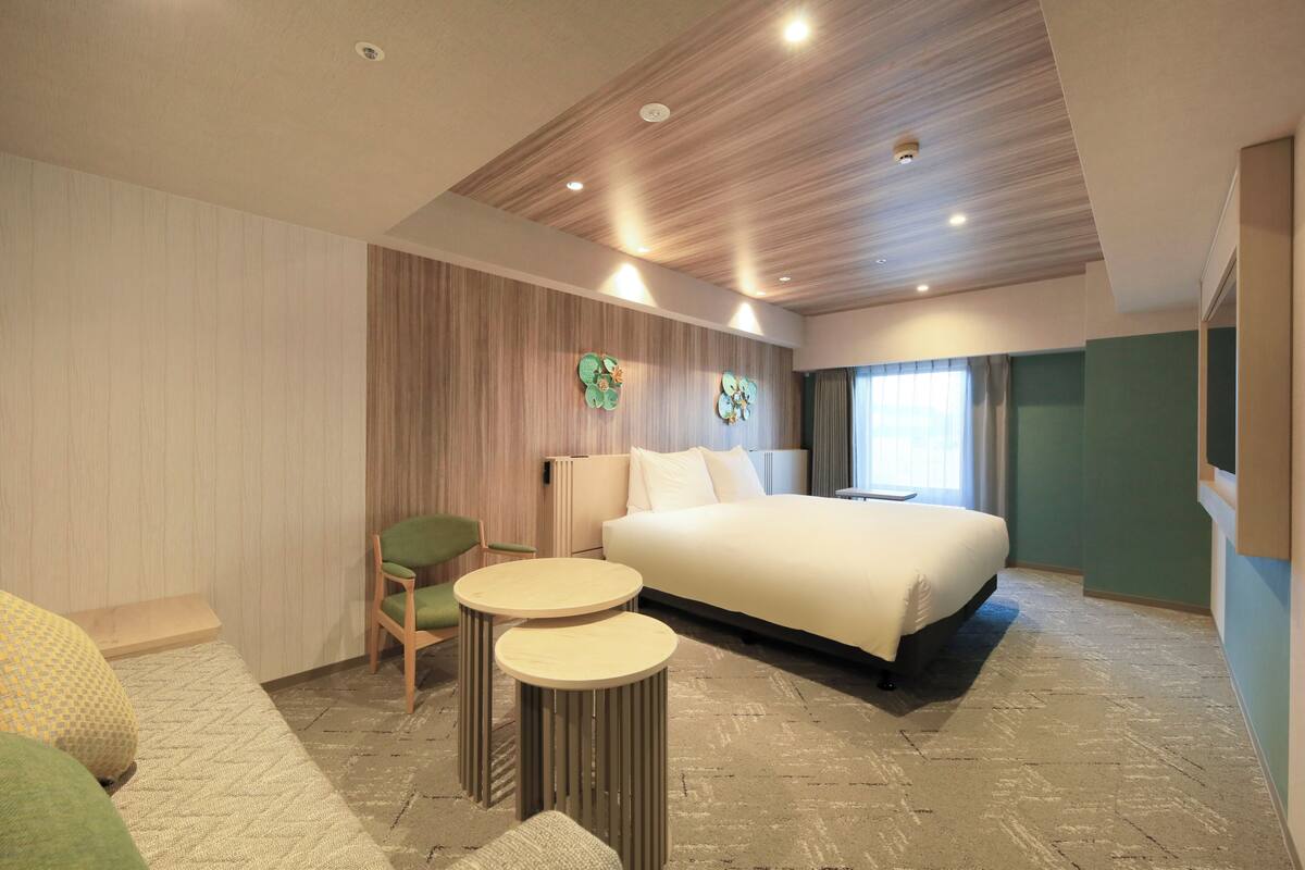A modern hotel room with a large bed, two round tables, a green chair, and a wooden ceiling.