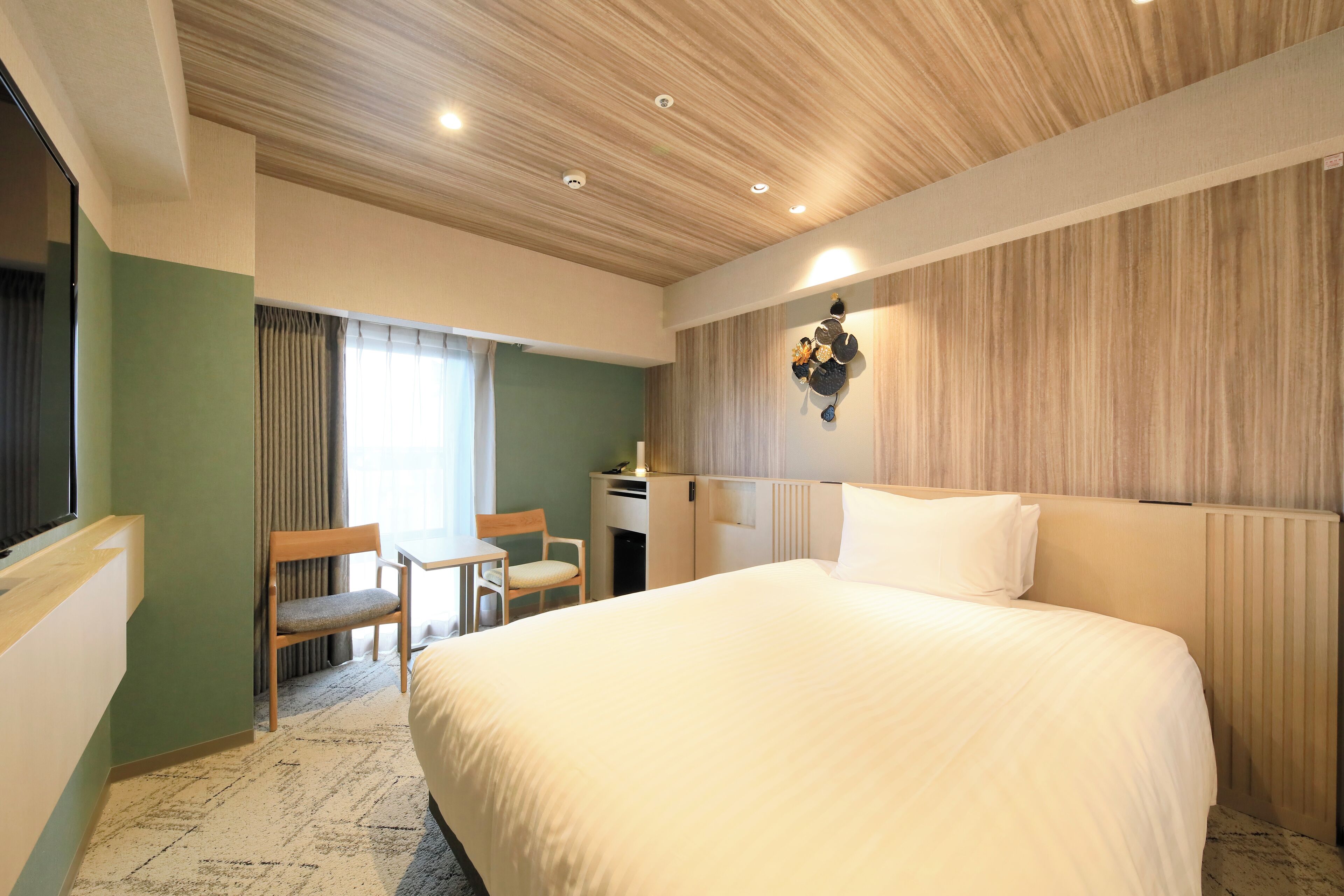 A hotel room with a large bed, a desk, two chairs, a window with curtains, and a wooden ceiling.