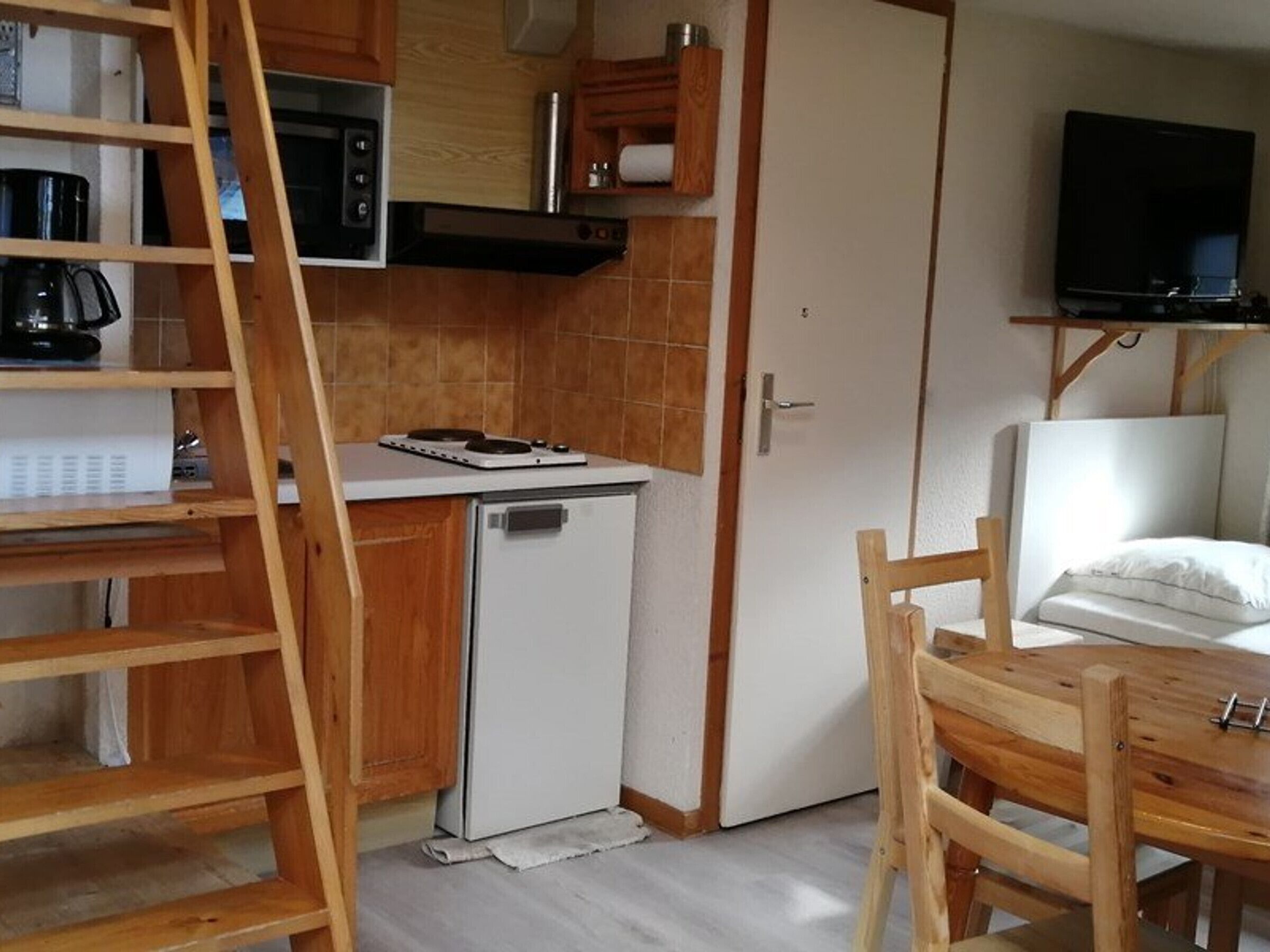 Une cuisine compacte équipée d’un four à micro-ondes, d’une cuisinière et d’un petit réfrigérateur. On y trouve une table en bois et une chaise, ainsi qu’un téléviseur fixé au mur.