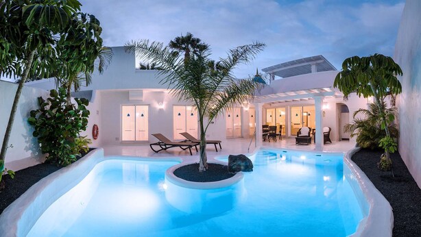 A pool area with lounge chairs and a dining area, surrounded by palm trees and a modern building. - Royal Villa 2 people Alegria (La Oliva)