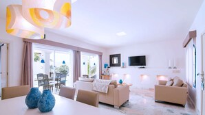 A modern living room with a sofa, two armchairs, a dining table with chairs, a TV, and a large window with curtains. - Royal Villa 2 people Alegria (La Oliva)