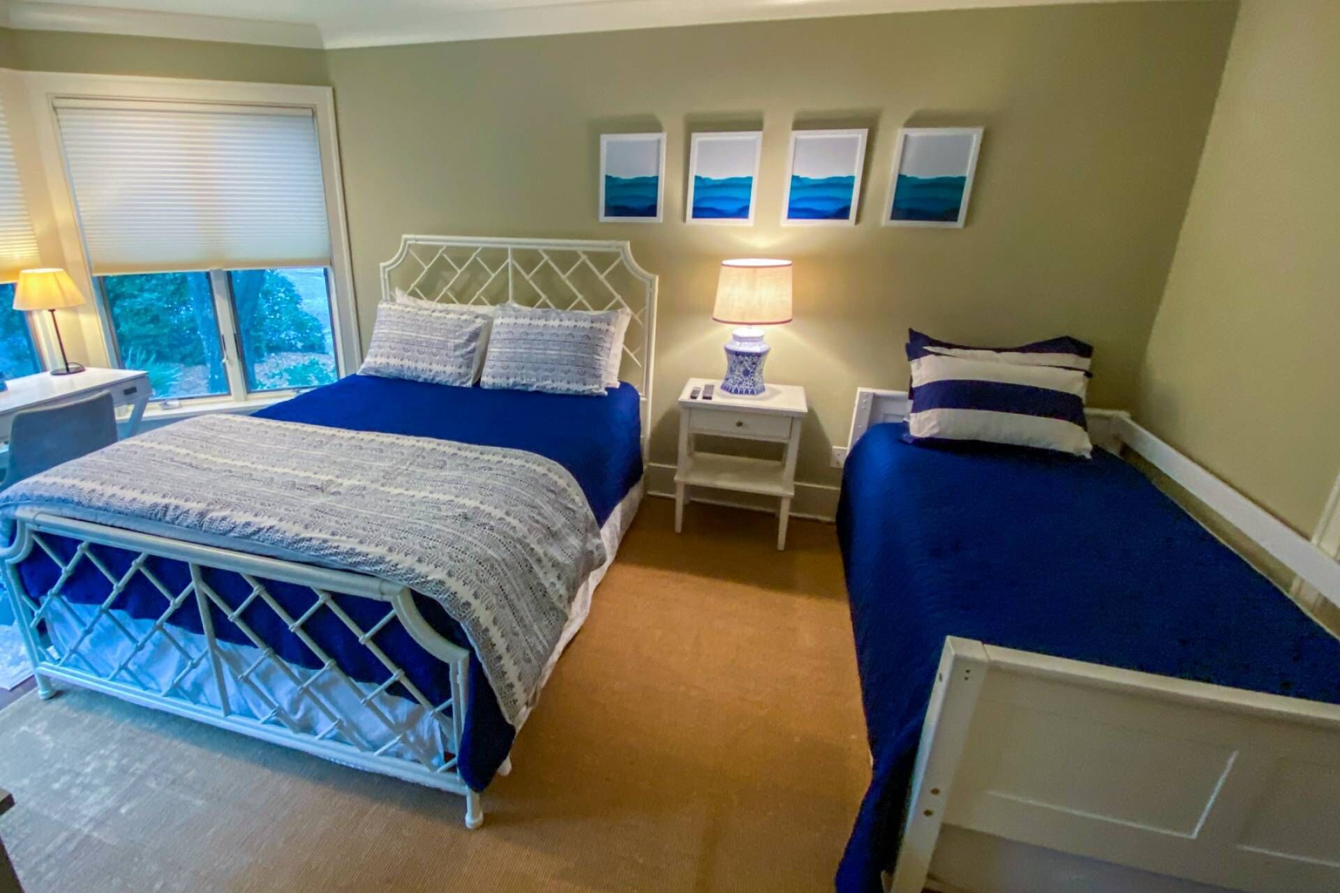 A bedroom with two beds, a nightstand with a lamp, and a window with blinds.