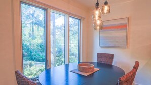 Una mesa de comedor redonda con un centro de mesa tejido, rodeada de sillas de mimbre, ventanas amplias con vista a un bosque y una lámpara colgante moderna.
