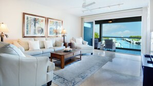 A modern living room with a sofa, coffee table, and artwork, leading to a balcony with a view of the sea and boats.