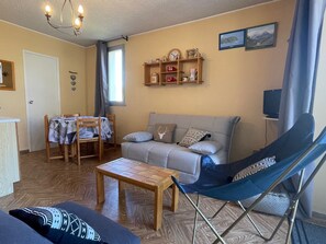 A cozy living room with a sofa, a dining table with chairs, a small wooden table, a blue folding chair, a chandelier, and a shelf with decorative items. - Studio in Sauze with balcony and garage (Enchastrayes)
