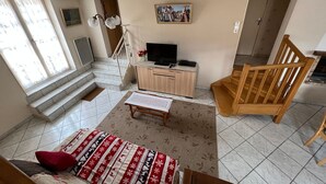 A living room with a sofa, a wooden chair, a TV on a wooden cabinet, a staircase, and a painting on the wall.