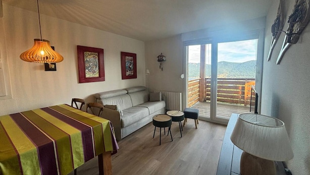 A cozy living room with a sofa, a dining table, and a balcony with a view. - 2-Bedroom Cabin Apartment with Parking, Lake View (Les Angles)