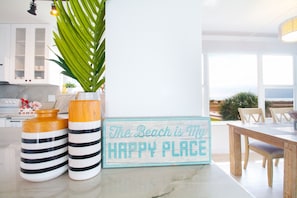 Une cuisine avec des meubles blancs, une plante en pot, des vases à rayures et un panneau sur lequel on peut lire : « La plage est mon endroit préféré. »
