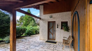 Eine überdachte Terrasse mit Steinboden, einer Holzbank, einer Tür und einem an der Wand befestigten Pflanzgefäß.