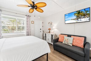 A bedroom with a bed, a sofa, a TV, and a ceiling fan. - Beachfront Studio at Tropic Terrace #49: Steps to sand, sunsets & Heated Pool (Treasure Island)