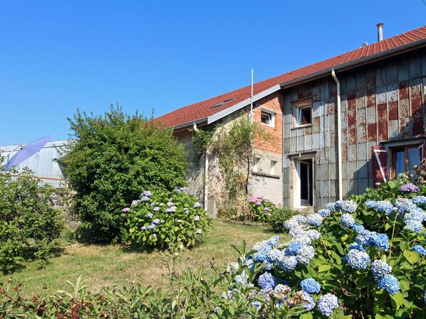 A rustic building with a red-tiled roof, surrounded by lush greenery and hydrangea flowers. - Charming Mountain Getaway with Fireplace, Near Gérardmer – Sleeps 10, Pet Friendly (Rehaupal)