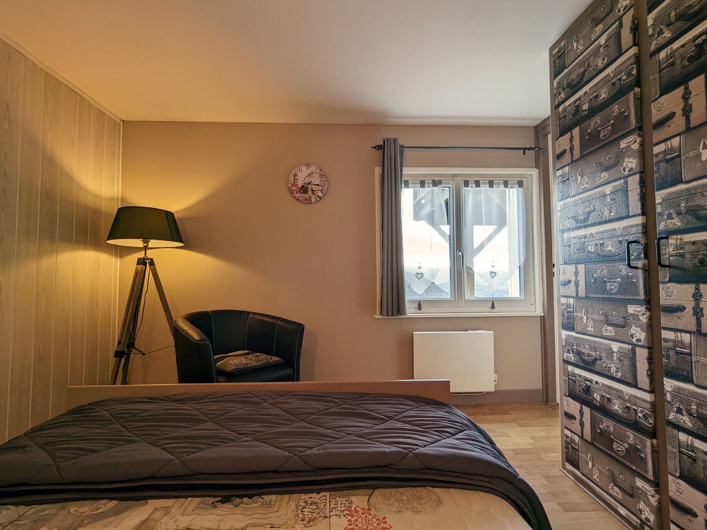 Un dormitorio con cama, una silla, una lámpara, una ventana con cortinas y una pared con maletas.