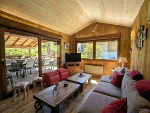 Een gezellig interieur van een chalet met houten plafond, een televisie, een bank, een eethoek en grote ramen.