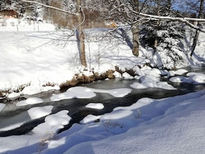 Un paysage enneigé avec un ruisseau gelé, des arbres et un bâtiment situé au loin.