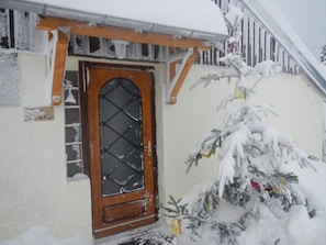 A snow-covered house with a wooden door and a small Christmas tree.