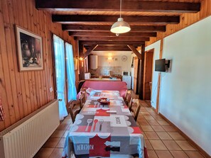 Uma sala de jantar com uma mesa comprida posta para várias pessoas, paredes de madeira, uma cozinha ao fundo e uma televisão fixada na parede.