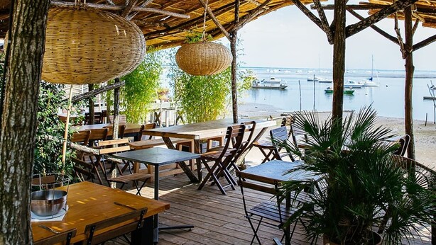 An outdoor dining area with wooden tables and chairs, hanging wicker baskets, and a view of the sea with boats.