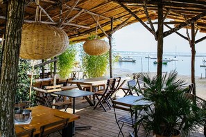 An outdoor dining area with wooden tables and chairs, hanging wicker baskets, and a view of the sea with boats. - Résidence Entre Océan/bassin - Beach Holiday Home (Lège-Cap-Ferret)