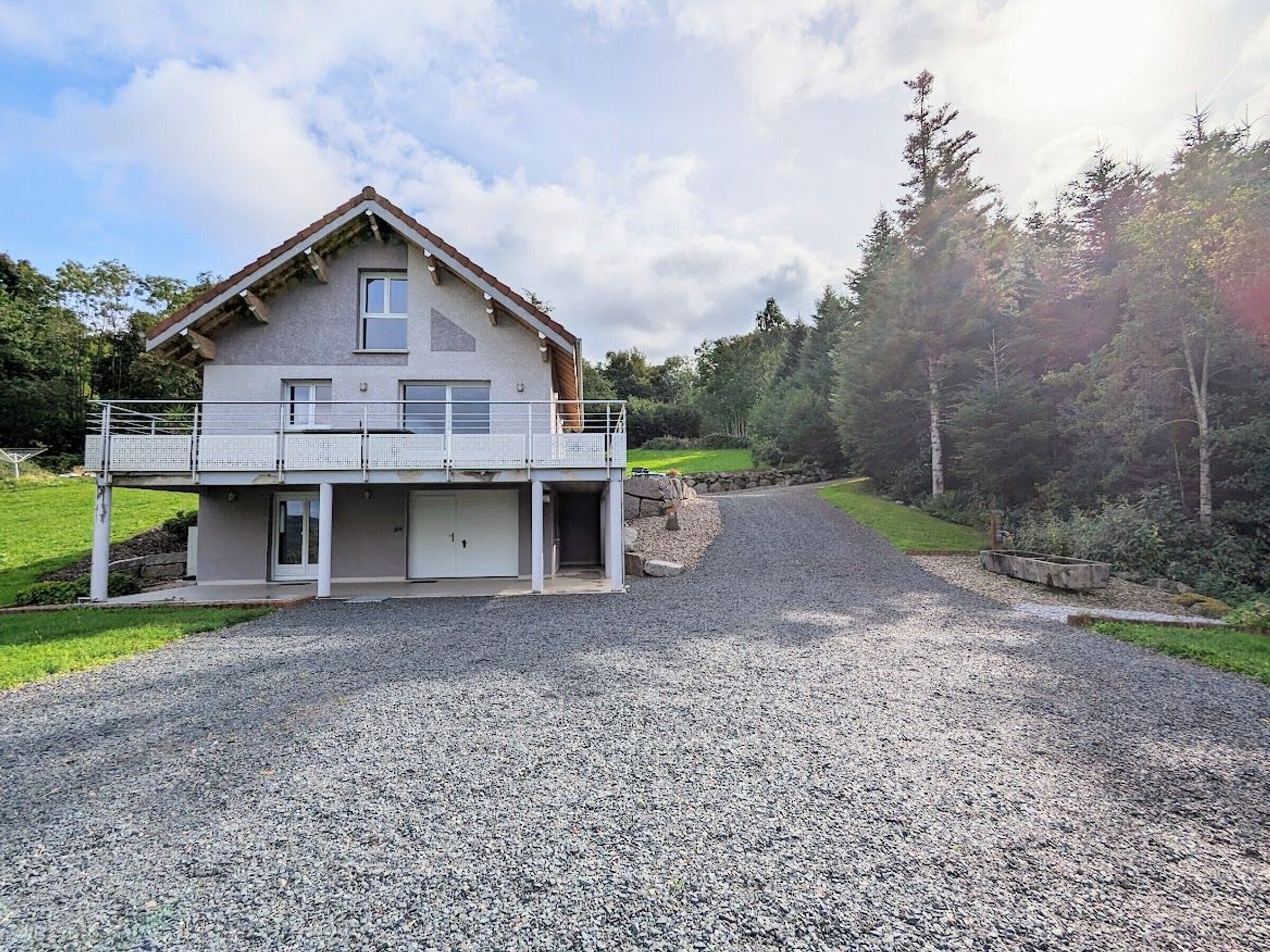 Ein zweigeschossiges Haus mit Balkon, Kiesauffahrt und bewaldeter Umgebung.