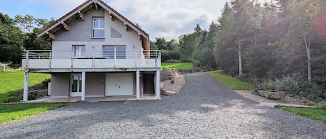 Ein zweigeschossiges Haus mit Balkon, Kiesauffahrt und bewaldeter Umgebung.