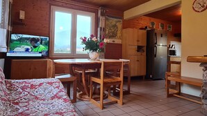 Un salon chaleureux avec un plafond en bois, un coin repas avec une table et des chaises en bois, une cuisine équipée d’un réfrigérateur et d’un four à micro-ondes, et un téléviseur diffusant un journal télévisé.