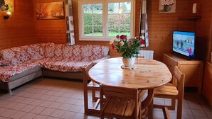 L’intérieur d’un chalet accueillant, avec un plancher en bois, une table ronde en bois entourée de chaises, un canapé, un téléviseur et une fenêtre agrémentée de rideaux.