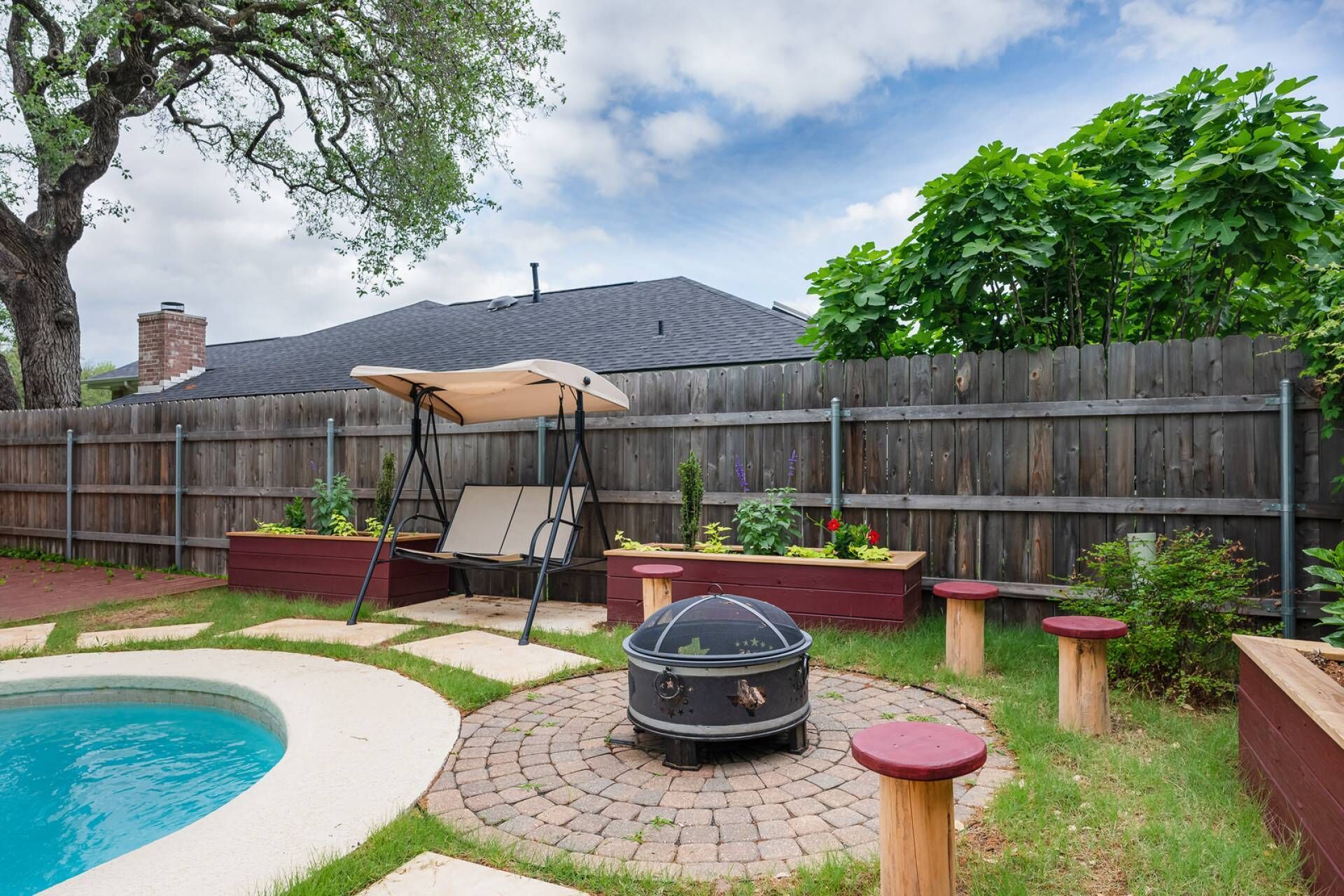 A backyard with a swimming pool, a swing chair, a fire pit, and wooden planters with plants.