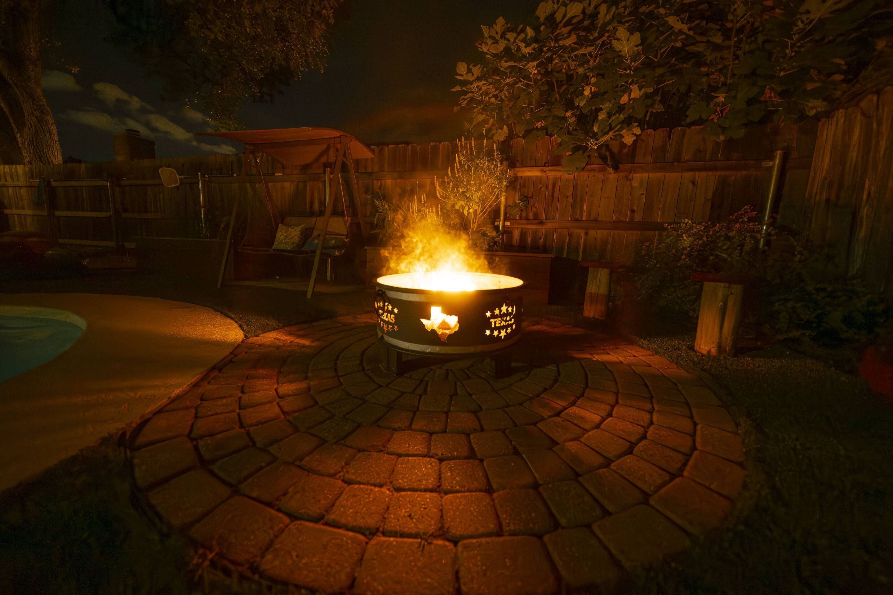 A circular outdoor fire pit with a metal base, surrounded by a paved area with a wooden fence and vegetation in the background.