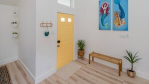 A hallway with a yellow door, two wall-mounted paintings of mermaids, a woven bench, and a potted plant. - Remodeled, Private Pool & Hot Tub, Gas BBQ, Xbox, Ping Pong, Foosball, Fully-equipped Kitchen (Round Rock)