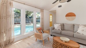 A living room with a sofa, a wooden side table, a wicker chair, and a large window overlooking a pool. - Remodeled, Private Pool & Hot Tub, Gas BBQ, Xbox, Ping Pong, Foosball, Fully-equipped Kitchen (Round Rock)