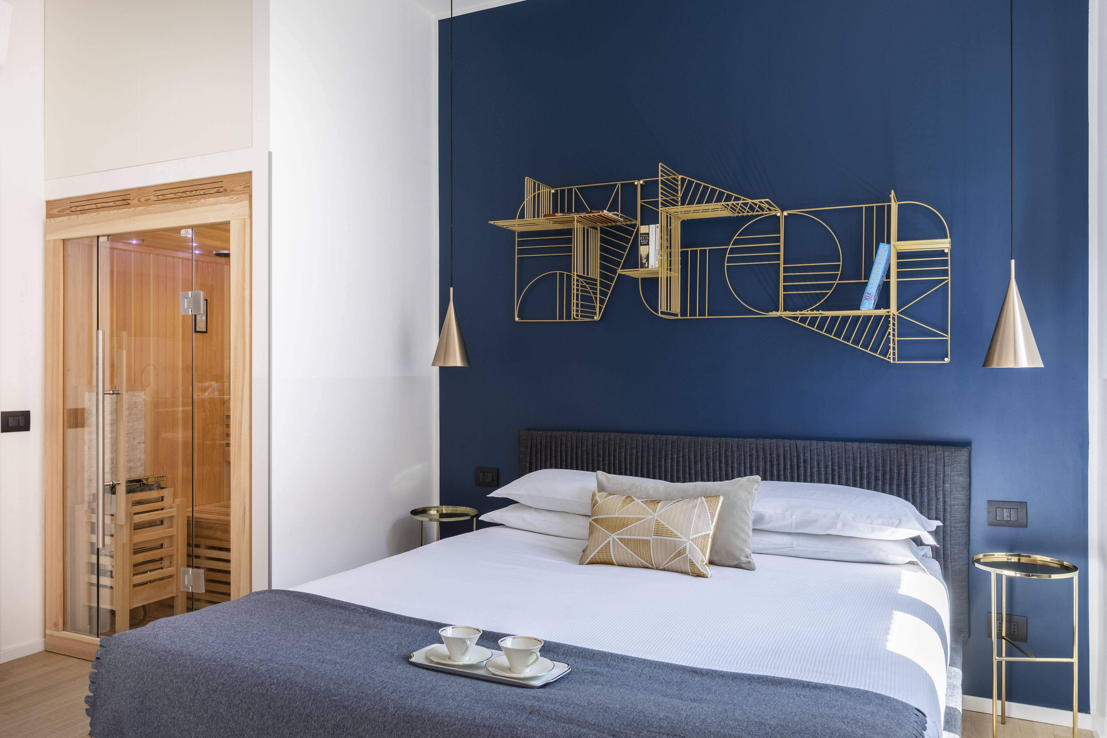 Une chambre d’hôtel moderne avec un grand lit, une décoration murale aux motifs géométriques, une armoire à portes en verre et une table de chevet avec une lampe.