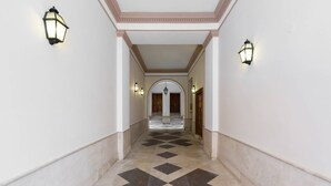 Um corredor com piso xadrez, paredes brancas e duas lanternas fixadas na parede.