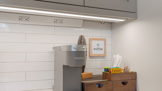 Una cocina moderna con una cafetera Keurig, un cartel enmarcado que dice 'coffee' y un mueble de madera con libros y utensilios de cocina.