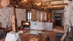 Un interior rústico con vigas de madera a la vista, pared de piedra, chimenea, zona de bar y área de descanso con una mesa de vidrio.