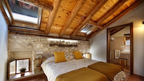 Un dormitorio con pared de piedra, vigas de madera, tragaluz, cama con dos almohadas y baño visible a través de una puerta abierta.