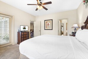A bedroom with a large bed, a ceiling fan, a dresser, a desk with a lamp, and a framed picture on the wall.