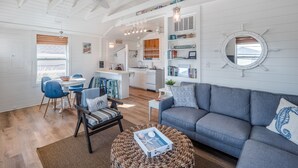 A spacious living room with a gray sofa, blue chairs, and a dining area with a table and chairs. - Cottage at Scotch Bonnet | Walk to the Beach! (Carolina Beach)