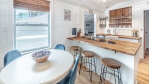 A modern kitchen with white cabinetry, a wooden countertop, and bar stools. There is a window with blinds, a bowl on the table, and a sign that reads "Seaside Bakery." - Cottage at Scotch Bonnet | Walk to the Beach! (Carolina Beach)
