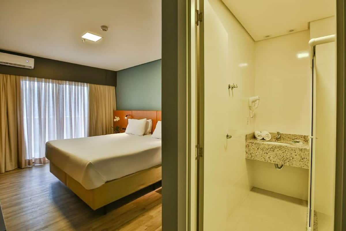 A hotel room with a bed, a bathroom with a shower, and a window with curtains.