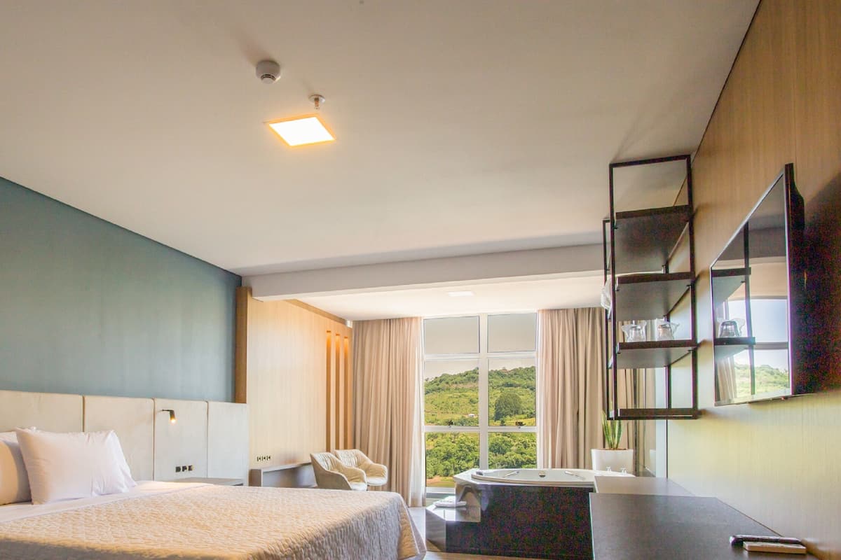A modern hotel room with a large bed, a desk, a chair, a bathtub, and a view of the outdoors.