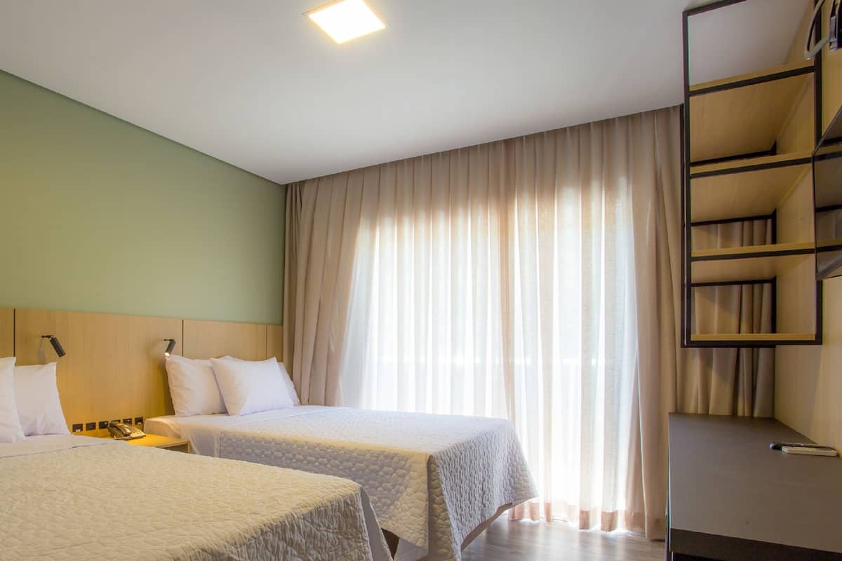 A hotel room with two beds, a wooden headboard, a bedside table with a lamp, a shelf with books, and a large window with curtains.